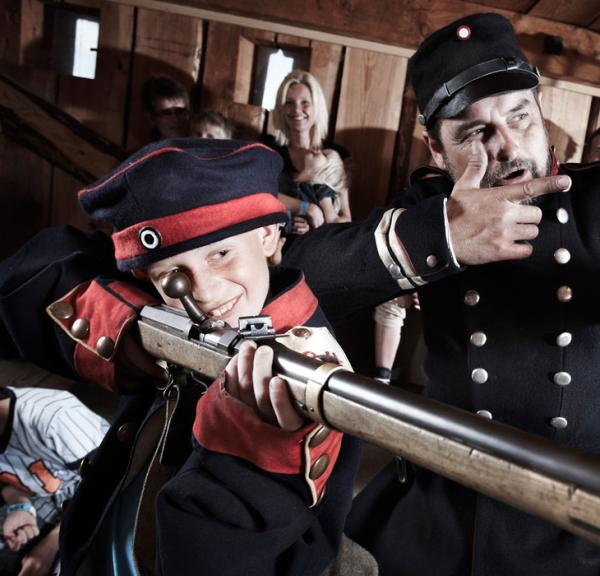 Medarbejder (formidler) og barn i uniform skyder med trægevær på Historiecenter Dybbøl Banke