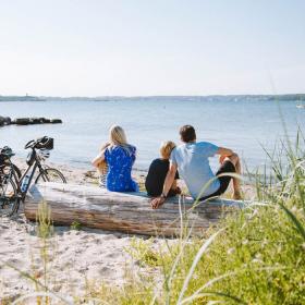 Familie sidder på stranden og kigger ud over vandet med cykler ved siden af